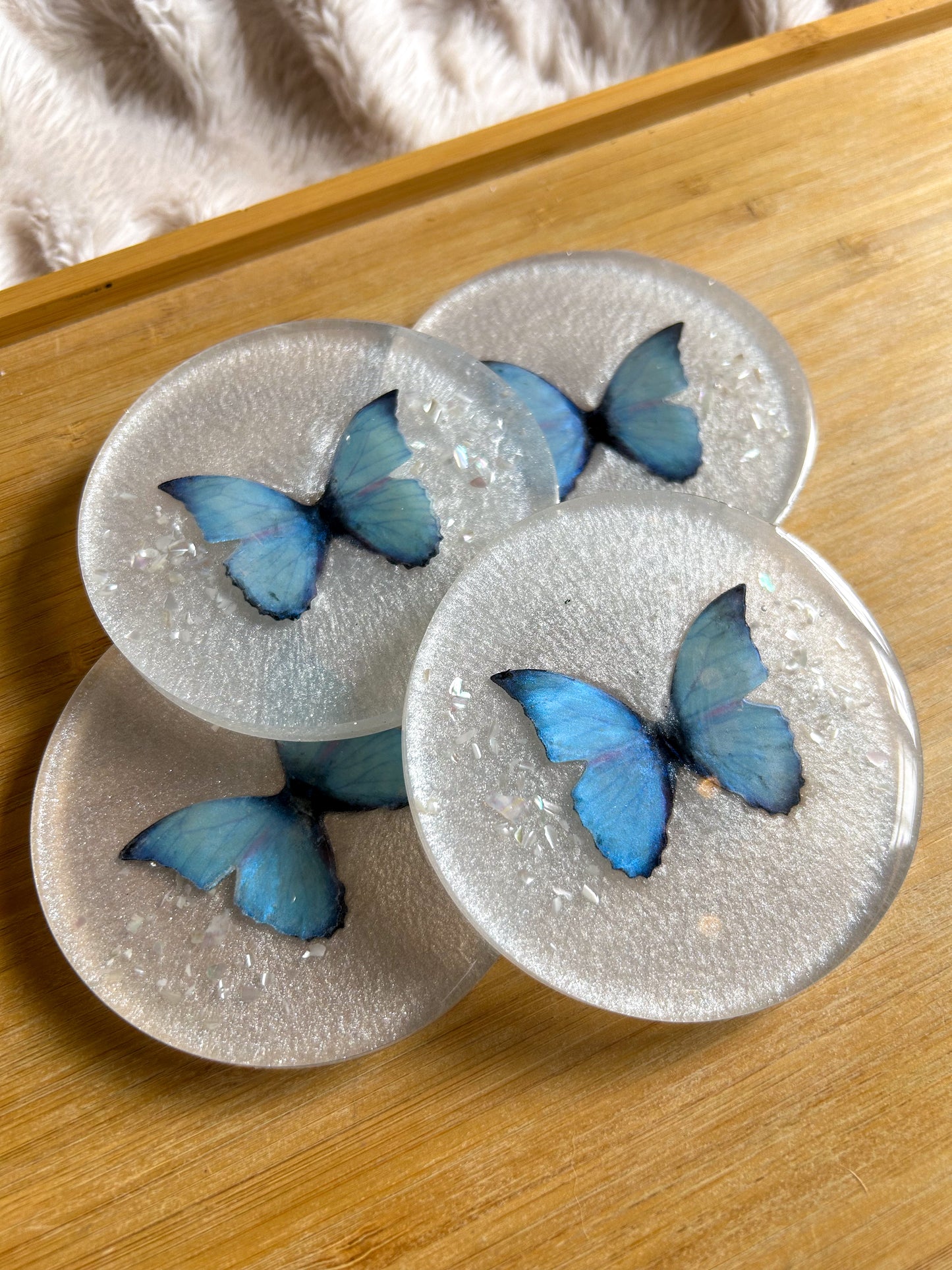Four round handmade resin coasters featuring a shimmering holographic blue monarch butterfly with delicate iridescent shell fragments. Elegant and artistic drink coasters with a glossy finish, perfect for home decor or gifting