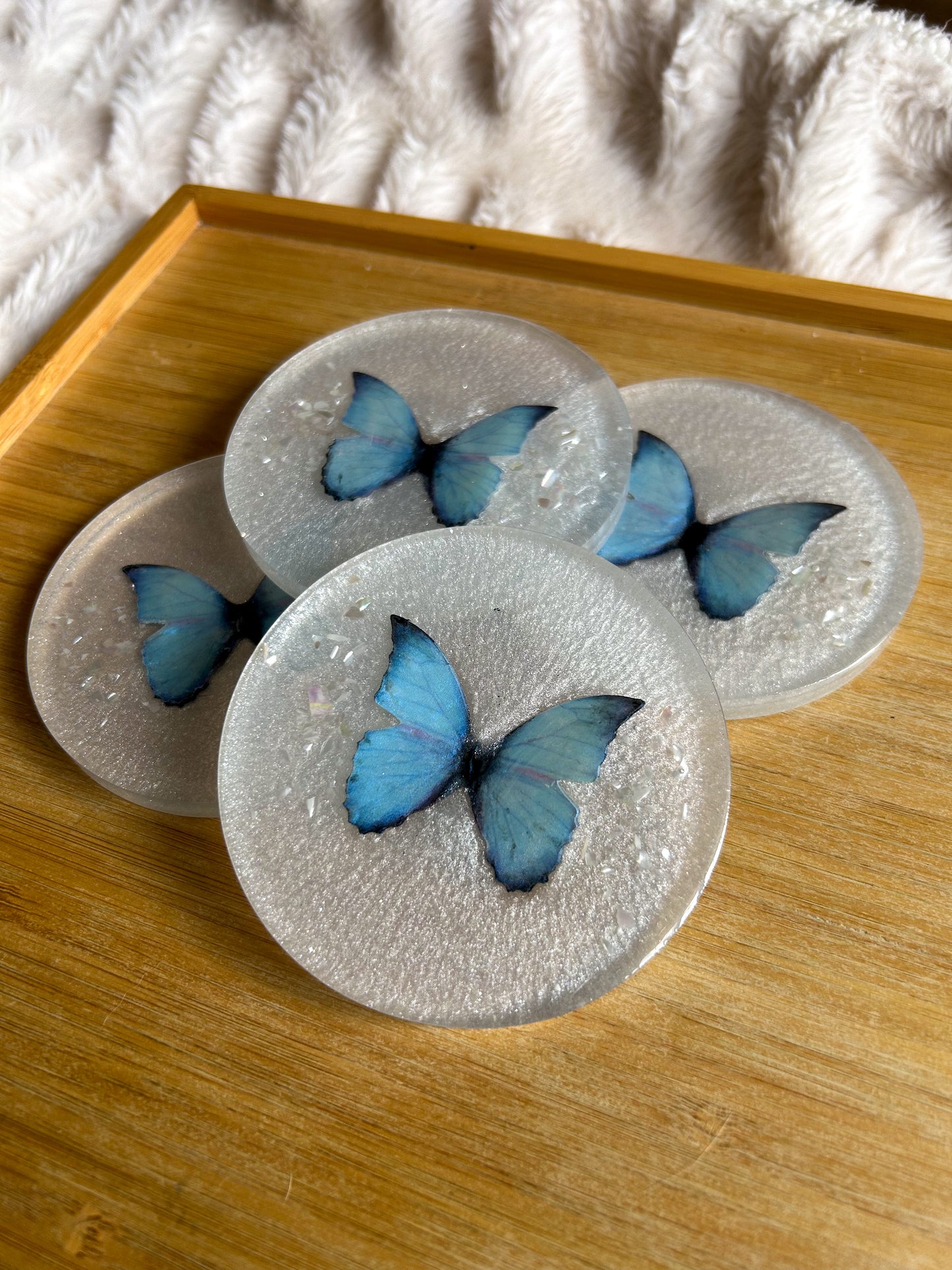 Four round handmade resin coasters featuring a shimmering holographic blue monarch butterfly with delicate iridescent shell fragments. Elegant and artistic drink coasters with a glossy finish, perfect for home decor or gifting