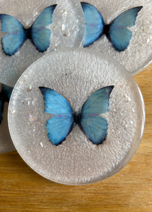 Four round handmade resin coasters featuring a shimmering holographic blue monarch butterfly with delicate iridescent shell fragments. Elegant and artistic drink coasters with a glossy finish, perfect for home decor or gifting