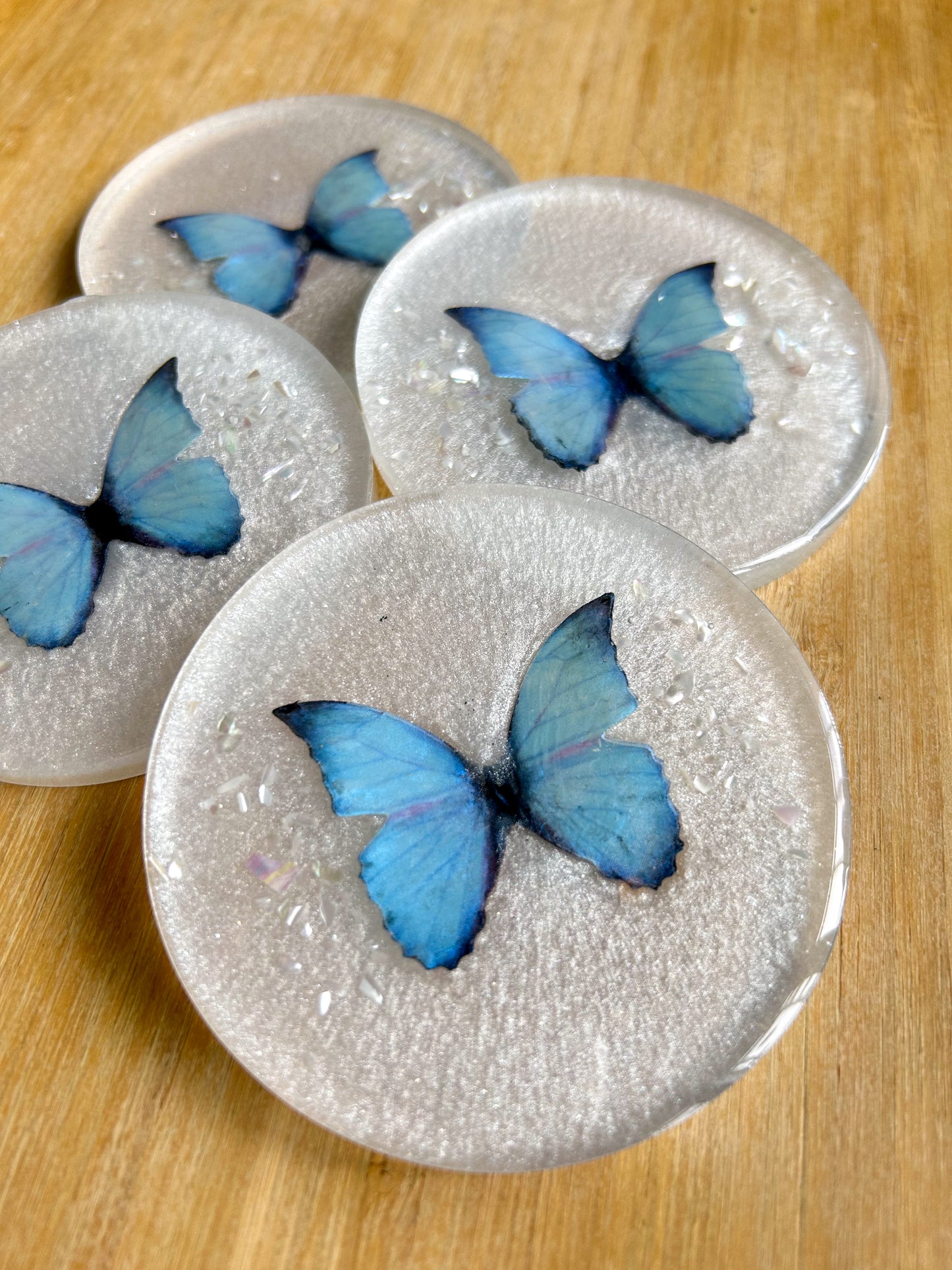 Four round handmade resin coasters featuring a shimmering holographic blue monarch butterfly with delicate iridescent shell fragments. Elegant and artistic drink coasters with a glossy finish, perfect for home decor or gifting