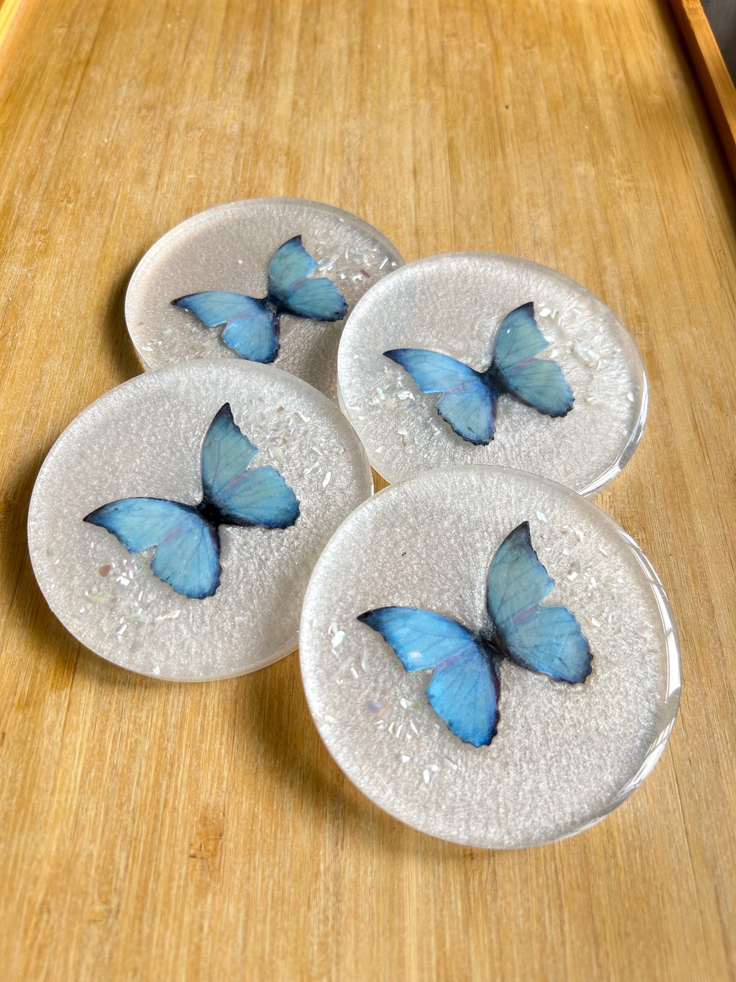 Four round handmade resin coasters featuring a shimmering holographic blue monarch butterfly with delicate iridescent shell fragments. Elegant and artistic drink coasters with a glossy finish, perfect for home decor or gifting