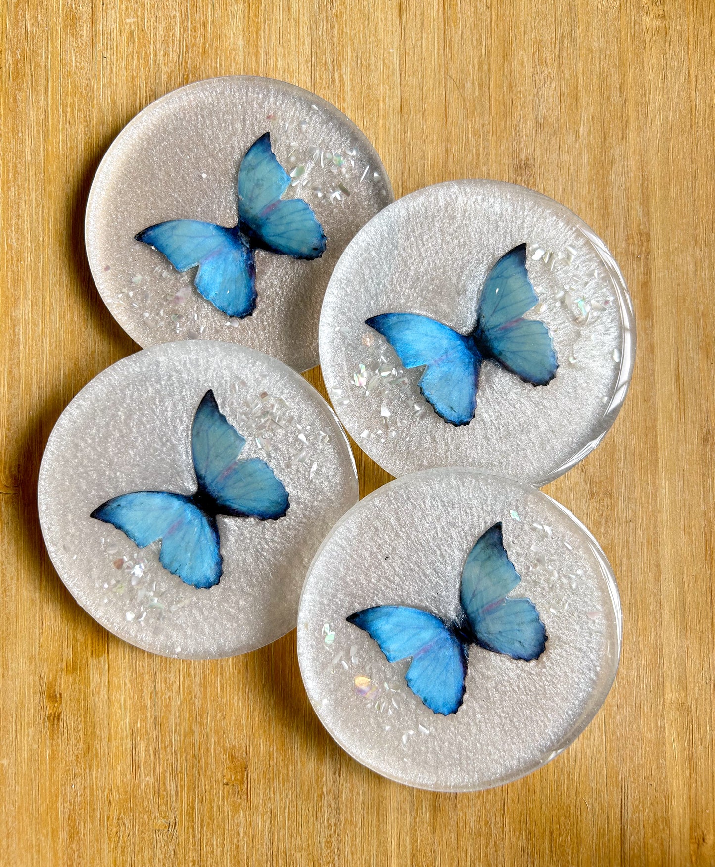 Four round handmade resin coasters featuring a shimmering holographic blue monarch butterfly with delicate iridescent shell fragments. Elegant and artistic drink coasters with a glossy finish, perfect for home decor or gifting