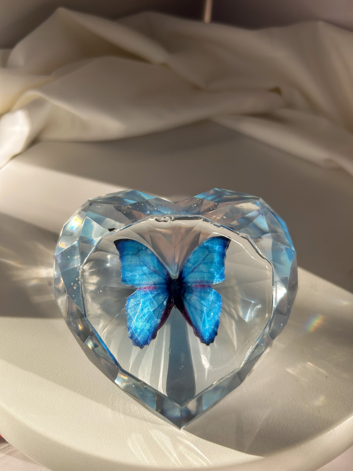 Heart-shaped resin paper holder featuring a faux blue monarch butterfly encased in clear resin. A nature-inspired handmade desk accessory perfect for home office decor or as a unique gift.