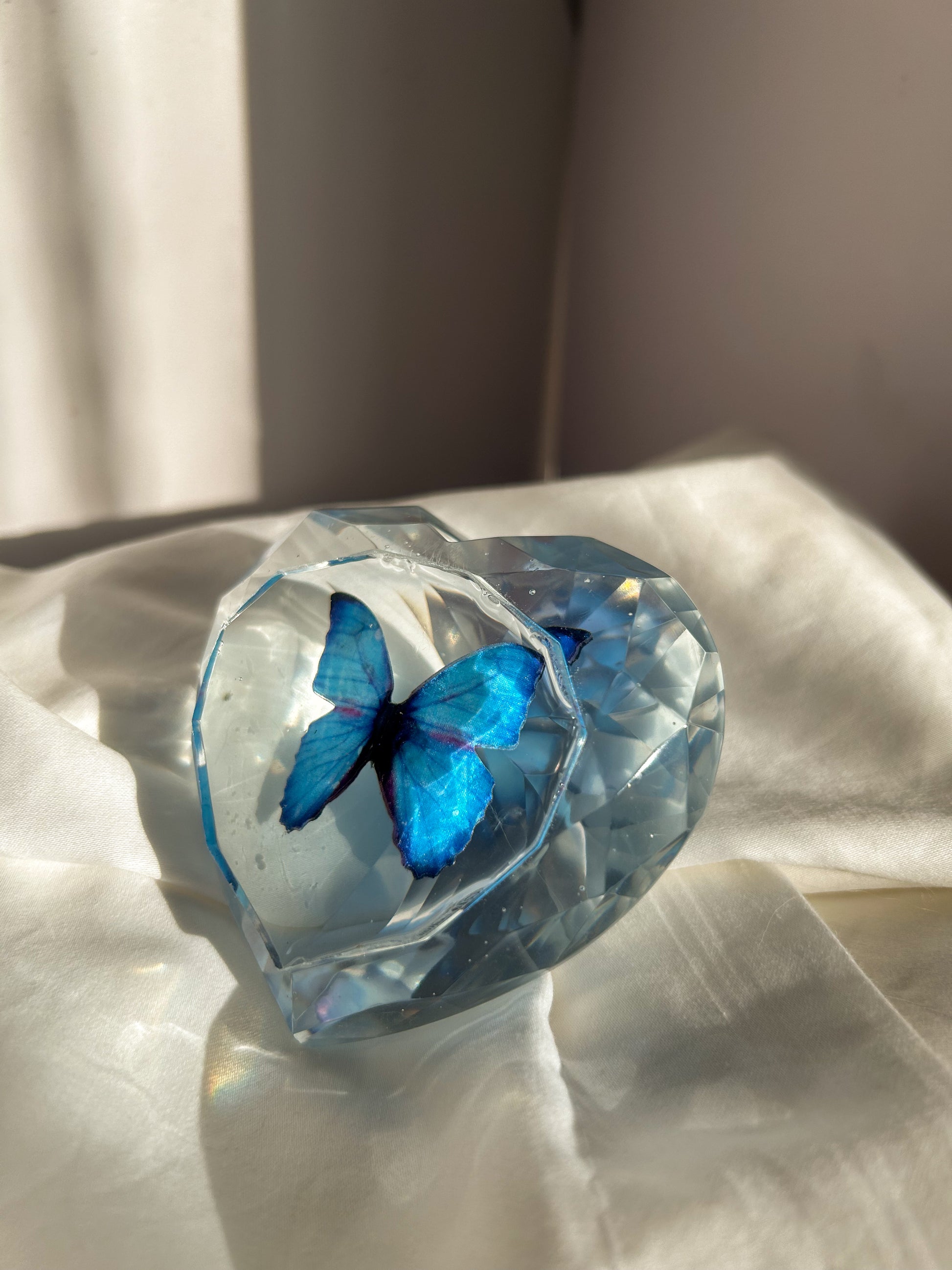 Heart-shaped resin paper holder featuring a faux blue monarch butterfly encased in clear resin. A nature-inspired handmade desk accessory perfect for home office decor or as a unique gift.